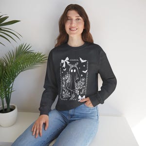 May include: A dark gray long-sleeve sweatshirt featuring a white graphic print of a ghost wearing a bow and patterned boots, holding a drink and a pumpkin bag. The design is framed by decorative borders with bows, bats, and floral elements.