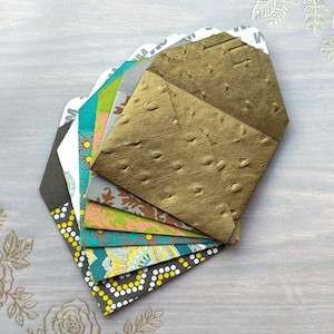 May include: A stack of colorful patterned envelopes with a gold embossed envelope on top. The envelopes are folded and stacked on a light gray surface.