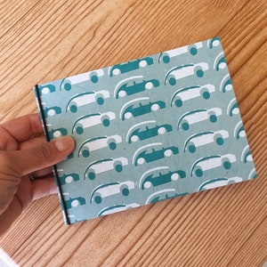 Puede incluir: Un pequeño cuaderno con un fondo turquesa y un patrón repetido de coches blancos y turquesas. El cuaderno está sujeto por una mano y tiene un cierre de banda elástica negra. La cubierta tiene una apariencia texturizada.