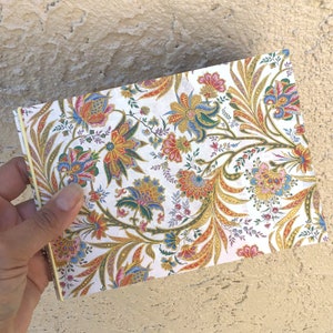 May include: A notebook with a floral pattern on a white background. The design includes colourful flowers in red, blue, and gold. The notebook is held in a hand, and the edges are visible.