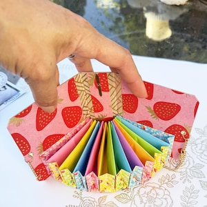 以下が含まれることがあります： カラフルなアコーディオンデザインの手作り紙財布。外側は、赤いイチゴのイラストが入ったピンク色の背景です。内側は折り紙の虹色です。金のチェーンが側面に付いています。