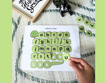 May include: A learning activity sheet with the text "I KNOW THE ALPHABET" and a green caterpillar design. The sheet has alphabet letters in green squares. A hand is holding the letter "U".