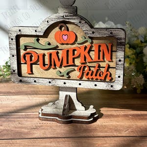 May include: A wooden sign with a distressed white paint finish reads "Pumpkin Patch" in orange letters. A small orange pumpkin with a heart is above the text. The sign is on a wooden stand.
