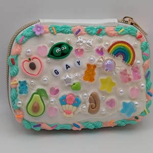 May include: A white, square-shaped zippered pouch decorated with colorful frosting, sprinkles, and various charms. The charms include a rainbow, gummy bears, hearts, stars, and the word "GAY".