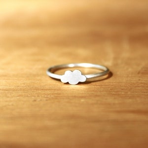 May include: A silver ring featuring a cloud-shaped design. The ring is displayed on a wooden surface.