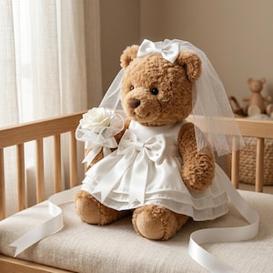 May include: A plush brown teddy bear wearing a white satin wedding dress with a bow and pearl accents. The bear has a white veil and holds a bouquet of white roses. White satin ribbon is draped around the bear.