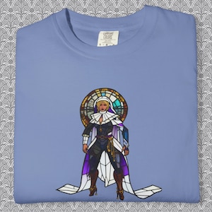 May include: A light blue Comfort Colors t-shirt featuring a stained-glass style illustration of a woman in a nun's habit. The artwork includes purple, white, and gold accents. The shirt has a tag that reads "Comfort Colors".