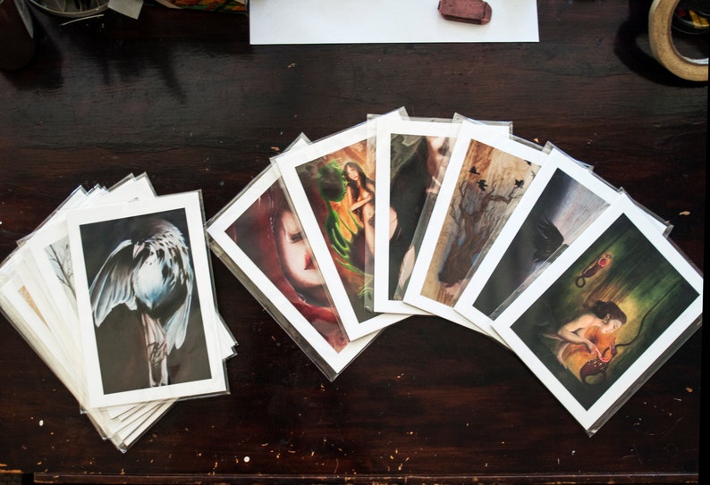 May include: A collection of art prints featuring various subjects, including a bird, a woman, and a tree. The prints are displayed in a fan shape on a wooden surface.