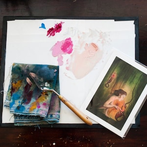 May include: A close-up of an artist's palette with a variety of paint colours, including pink, red, and blue. A painting knife and a piece of fabric with paint splatters are also visible. A framed print of a woman holding a red object is in the background.