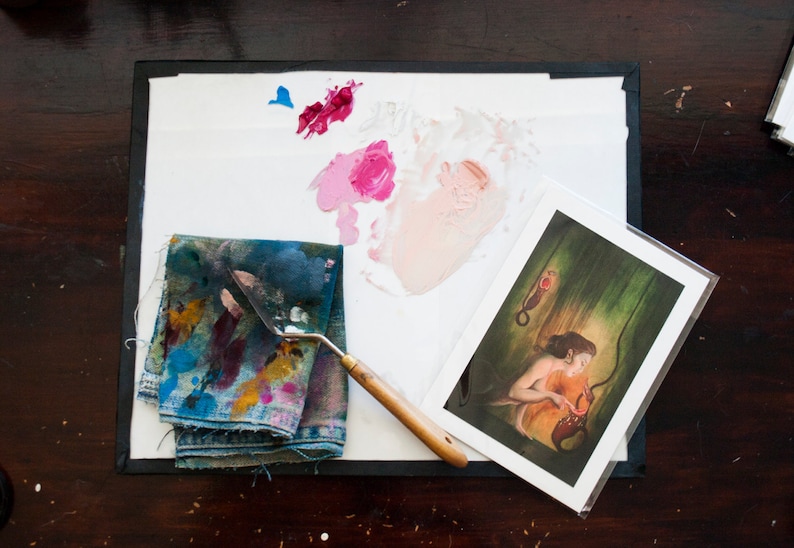 May include: A close-up of an artist's palette with a variety of paint colours, including pink, red, and blue. A painting knife and a piece of fabric with paint splatters are also visible. A framed print of a woman holding a red object is in the background.