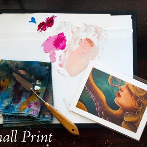 May include: A small print of a woman with an octopus tentacle wrapped around her neck. The print is on white cardstock and has a white border. The print is surrounded by a white palette with paint splatters and a blue cloth with paint splatters.