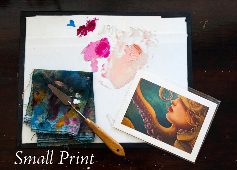 May include: A small print of a woman with an octopus tentacle wrapped around her neck. The print is on white cardstock and has a white border. The print is surrounded by a white palette with paint splatters and a blue cloth with paint splatters.