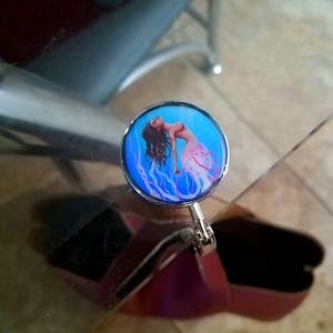 May include: A silver-toned metal clip with a round, blue and pink image of a mermaid with long, flowing hair. The mermaid is depicted with a pink tail and is surrounded by blue and white swirls.