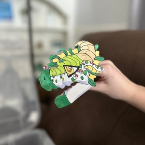 Op de afbeelding: Een handgeschilderde drakenkop-uitsparing met groene, gele en witte details. De draak heeft grote gele ogen en kleine paddenstoeldetails. De uitsparing wordt door een hand vastgehouden.