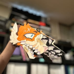Op de afbeelding: Een handgemaakte dierenkop van papier in oranje, wit en zwart. De kop heeft een cartoonachtig ontwerp met blauwe ogen en een gedetailleerde, gelaagde constructie. De achtergrond is wazig, wat wijst op een binnenomgeving.