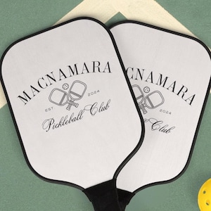 Könnte beinhalten: Zwei weiße Pickleball-Schläger mit schwarzen Griffen und schwarzer Beschriftung. Die Schläger tragen den Text "MacNamara Pickleball Club Est. 2024". Zwei gelbe Pickleball-Bälle befinden sich im Vordergrund.