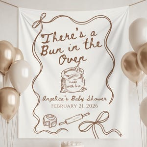 Peut inclure: Une bannière blanche avec l'inscription marron "There's a Bun in the Oven" sur le thème d'une baby shower. La bannière comprend des illustrations d'un sac de farine, d'un rouleau à pâtisserie et d'une pâtisserie. Des ballons dorés, blancs et transparents ornent la scène.