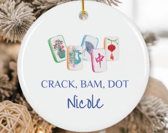 Könnte beinhalten: Runder weißer Keramik-Weihnachtsschmuck mit illustrierten Mahjong-Kacheln. Die Kacheln zeigen Blumen-, Pfauen- und Laternenmotive. Der Text "CRACK, BAM, DOT" und der Name "Nicole" sind in blauer Schrift geschrieben. Ein goldener Faden ist befestigt.