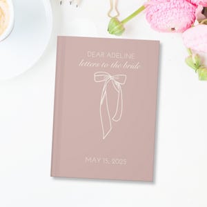 May include: A blush pink journal with the text "Dear Adeline letters to the bride" and a white ribbon bow. The date "May 15, 2025" is printed below the bow.