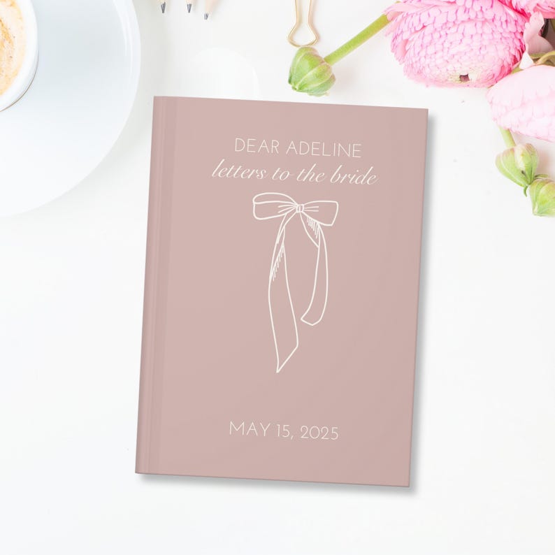 May include: A blush pink journal with the text "Dear Adeline letters to the bride" and a white ribbon bow. The date "May 15, 2025" is printed below the bow.