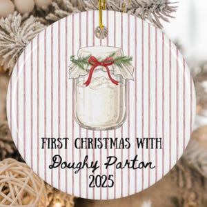 May include: A round Christmas ornament with a jar illustration. The jar is filled with a white substance and decorated with a red bow and greenery. The ornament has a red and white striped background and text that reads "FIRST CHRISTMAS WITH Doughy Parton 2025."