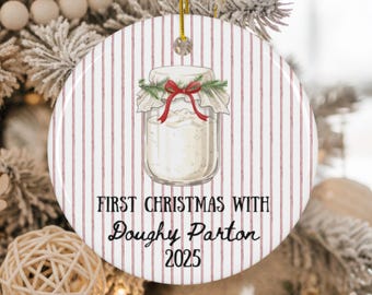 May include: A round Christmas ornament with a jar illustration. The jar is filled with a white substance and decorated with a red bow and greenery. The ornament has a red and white striped background and text that reads "FIRST CHRISTMAS WITH Doughy Parton 2025."