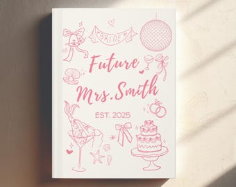 May include: A white notebook with pink illustrations and text. The cover features a banner that says "Bride", a disco ball, a mermaid in a martini glass, a cake, and the words "Future Mrs. Smith EST. 2025".