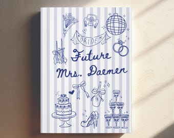 Op de afbeelding: Een rechthoekig notitieboek met een lichtblauw en wit verticaal gestreepte kaft, voorzien van donkerblauwe lijntekeningen van een bruidstaart, champagnetoren, discobal, ringen en de tekst "Future Mrs. Daemen" en "BRIDE".