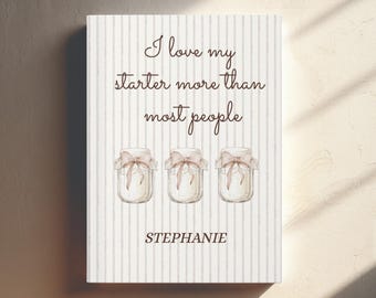 Könnte beinhalten: Ein Buchcover mit dem Text "I love my starter more than most people" in brauner Schrift. Darunter sind drei Gläser mit Deckeln, die mit Schleifen gebunden sind. Der Name "Stephanie" ist unten aufgedruckt. Der Hintergrund hat ein vertikales Streifenmuster.
