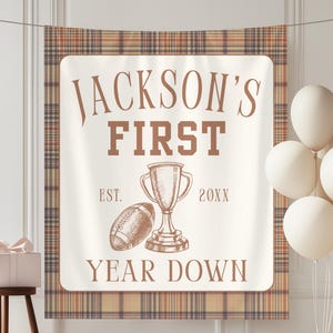 May include: A rectangular fabric banner featuring a brown plaid border and a cream center with the text JACKSON'S FIRST YEAR DOWN. The design includes a sketch of a football and a trophy, with the text EST. 20XX flanking the central trophy graphic.