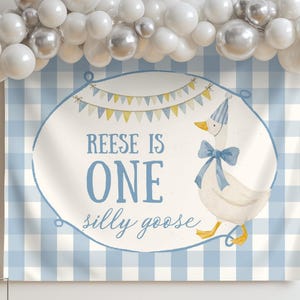 May include: A blue and white gingham backdrop with a banner that reads "Reese is One silly goose." The banner features a cartoon goose wearing a party hat and bow. A white cake and balloon arch are also visible.