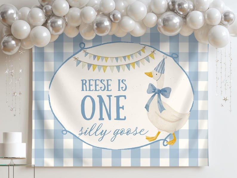 May include: A blue and white gingham backdrop with a banner that reads "Reese is One silly goose." The banner features a cartoon goose wearing a party hat and bow. A white cake and balloon arch are also visible.