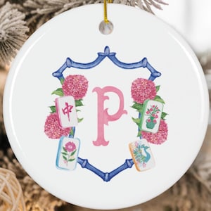 Puede incluir: Adorno navideño de cerámica blanca con una letra 'P' rosa en el centro, rodeada por un marco azul y flores rosas. Cuatro fichas de Mahjong están colocadas alrededor de la letra. Una cuerda dorada está unida para colgar.