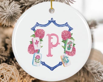 Puede incluir: Adorno navideño de cerámica blanca con una letra 'P' rosa en el centro, rodeada por un marco azul y flores rosas. Cuatro fichas de Mahjong están colocadas alrededor de la letra. Una cuerda dorada está unida para colgar.