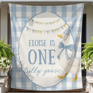May include: A light blue and white gingham banner with an oval design featuring a goose wearing a party hat and bow. Text reads "Eloise is One silly goose". The banner is hanging on a porch.