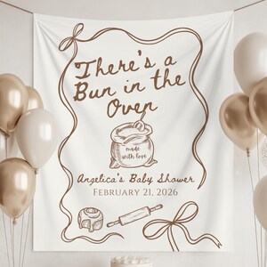 May include: A white banner with brown text and illustrations for a baby shower. The banner reads "There's a Bun in the Oven" with the name "Angelica's Baby Shower" and the date "FEBRUARY 21, 2026". Balloons in gold and white are on either side.