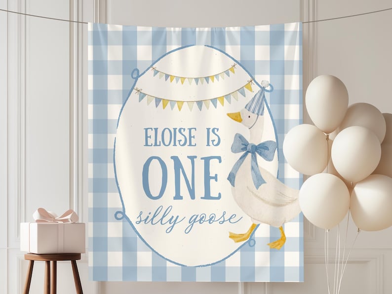 May include: A blue and white gingham backdrop with a banner that reads "Eloise is One, a silly goose." A cartoon goose wearing a party hat and bow tie is featured. White balloons and a gift box complete the scene, perfect for a first birthday.