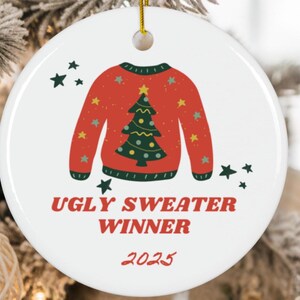 May include: A white, round Christmas ornament with a red sweater design featuring a green Christmas tree. The text "UGLY SWEATER WINNER 2025" is printed in red. Gold string for hanging.