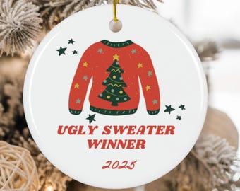 以下が含まれることがあります： 赤いセーターのデザインと緑のクリスマスツリーが特徴の、白い丸いクリスマスオーナメント。「UGLY SWEATER WINNER 2025」の文字が赤でプリントされています。吊り下げ用の金色の紐。