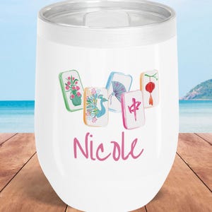 May include: White insulated wine tumbler with a silver rim, featuring colorful Mahjong tile illustrations and the name "Nicole" in pink script. The background shows a beach scene with blue water and sky.