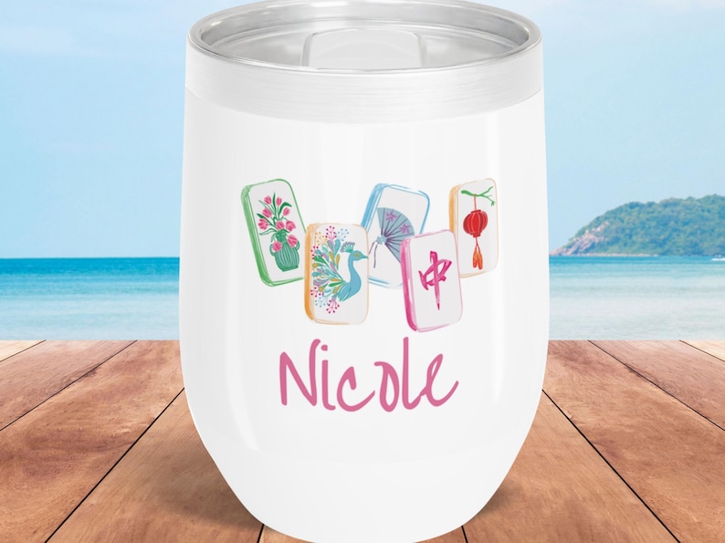 May include: White insulated wine tumbler with a silver rim, featuring colorful Mahjong tile illustrations and the name "Nicole" in pink script. The background shows a beach scene with blue water and sky.