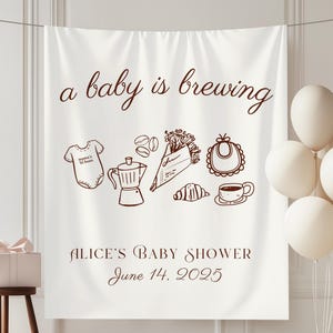 May include: A white baby shower backdrop with the text "a baby is brewing" and illustrations of baby items and coffee. The backdrop also includes the text "ALICE'S BABY SHOWER June 14, 2025". White balloons are on the right.
