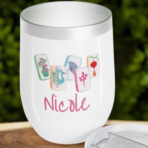 May include: White stemless wine tumbler with a silver rim, featuring a watercolor design of Mahjong tiles and the name "Nicole" in pink script. The tumbler has a lid.
