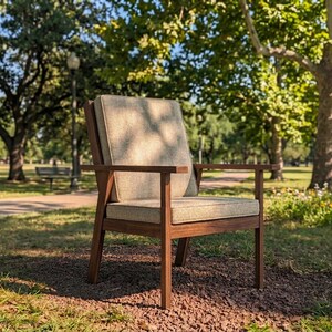 Puede incluir: Un sill&oacute;n de madera con un coj&iacute;n y respaldo de color claro se encuentra sobre un lecho de peque&ntilde;as rocas y hierba. La silla tiene un marco y reposabrazos de color marr&oacute;n oscuro. El fondo presenta un parque con &aacute;rboles y un sendero.