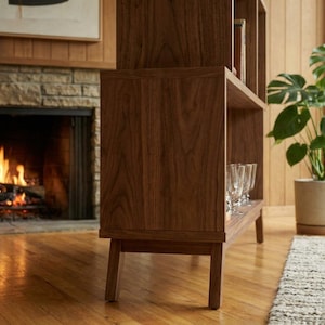 May include: A mid-century modern style wooden bookshelf with multiple shelves. The bookshelf is a rich brown color and has a rectangular shape. The shelves are empty except for a few wine glasses. A fireplace with a burning fire is in the background.