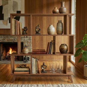 May include: A mid-century modern wooden bookshelf with multiple shelves, displaying books, vases, and decorative objects. The warm wood tones complement the room's decor, with a fireplace and a potted plant in the background.