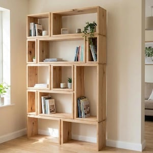 May include: A light-colored wooden bookshelf with multiple cubbyhole shelves of varying sizes. The shelves are filled with books, small plants, and decorative items. The bookshelf is against a light-colored wall.