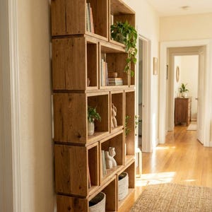 May include: A tall, wooden bookshelf with a stacked, open-cube design. The shelves hold books, plants, and decorative items. The natural wood finish complements the neutral-toned interior. The bookshelf is placed against a light-colored wall.