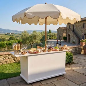 Op de afbeelding: Een witte buitenbar met een crèmekleurige geschulpte parasol. De bar is gedekt met wijnglazen, kaas, brood en fruit. De bar staat op een stenen terras met een schilderachtige achtergrond van een wijngaard en een stenen gebouw.