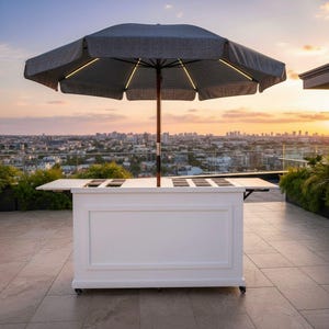 May include: A white outdoor bar with a grey patterned umbrella. The bar has a rectangular design with recessed compartments on top. The umbrella is open, with lights along the edges. The bar is on wheels, set against a city skyline at sunset.